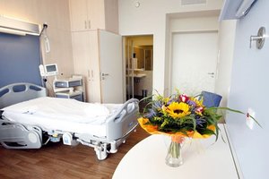 Patientenzimmer mit Pflegebett, Tisch mit Blumenstrauß und Stühlen